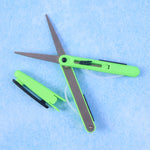 Green pen-shaped scissors with a black safety cutter are displayed on a light blue textured surface.