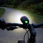 A person's hand on bicycle handlebars with the red and black headlight shining a bright beam on a paved path.