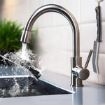 A black and gray crevice cleaning brush is being rinsed under a stainless steel kitchen faucet with water splashing.