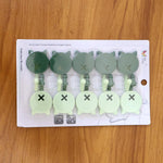 Ten cat-shaped wall hooks, five dark green and five light green, packaged in clear plastic on a wooden surface.