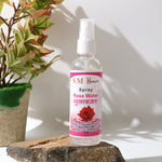 A clear bottle of SM Beauty rose water spray with a pink label stands on a rock next to a green plant.