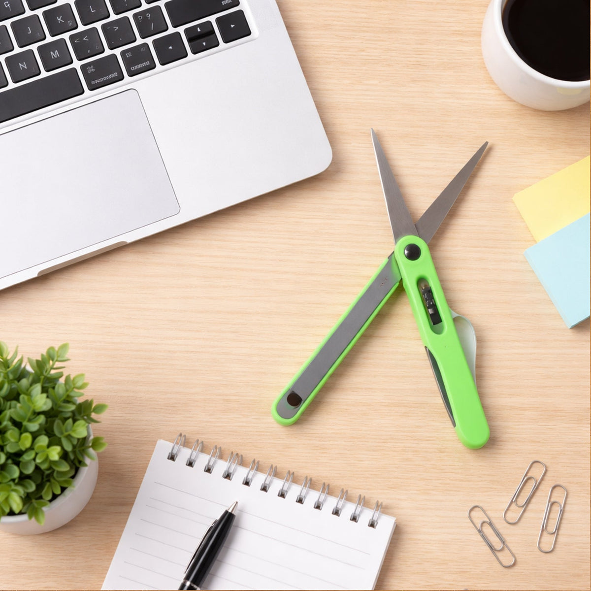 Green pen-shaped scissors and a black safety cutter lie on a wooden desk next to a laptop, notebook, and coffee.