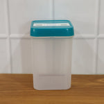 Empty clear plastic food storage container with a teal lid on a wooden surface against a white tiled wall.