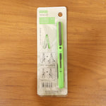 The green pen-shaped safety cutter is displayed in its retail packaging on a wooden surface.