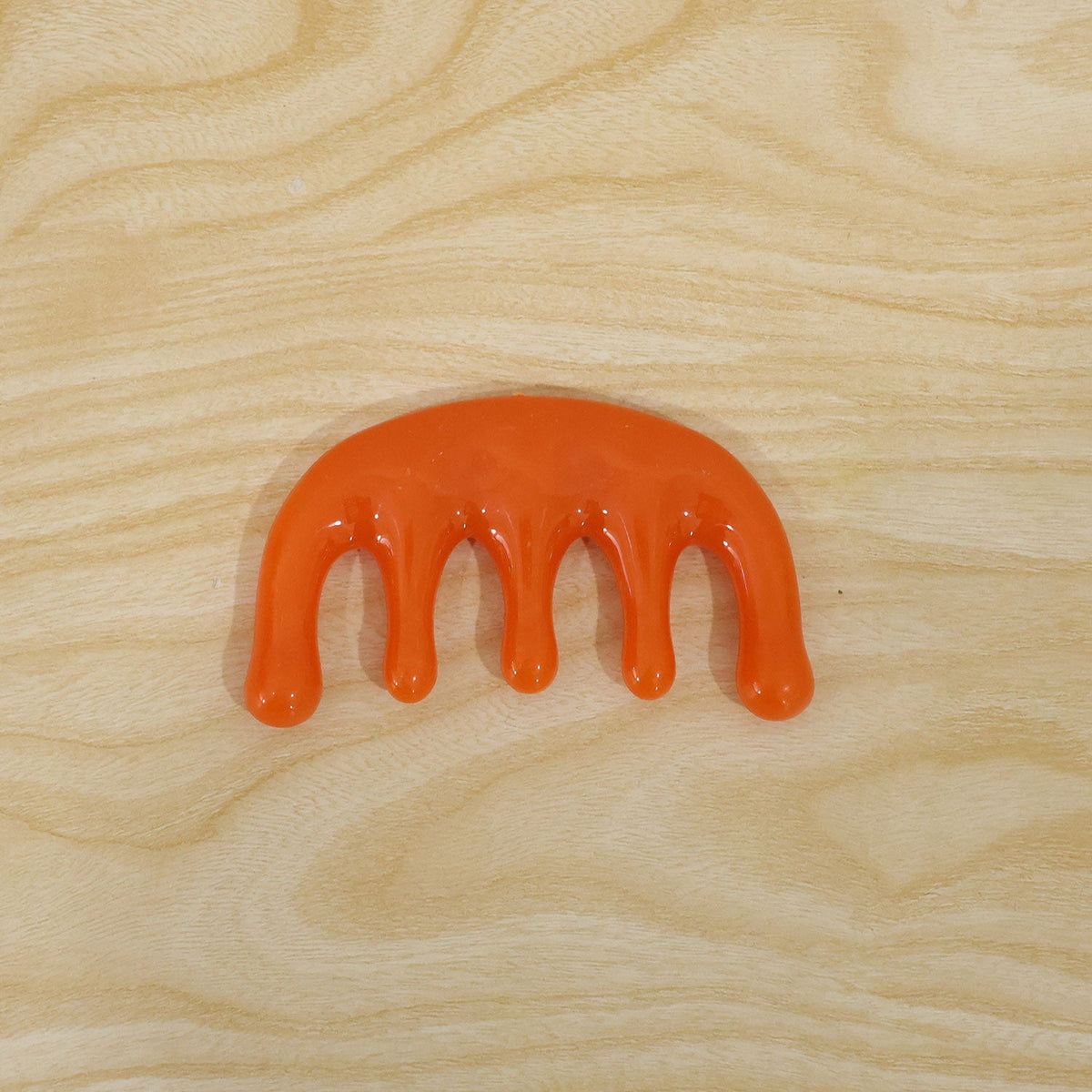An orange plastic scalp massager with five rounded prongs lies flat on a light wood grain surface.