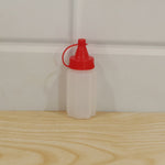 A small, empty, white ribbed plastic squeeze bottle with a red cap sits on a light wooden surface against a white wall.