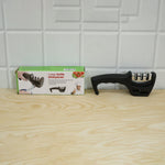 A black three-stage knife sharpener and its product box are placed side-by-side on a light wooden surface.