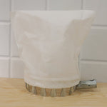 The white collection bag and metal prongs of the fruit picker are on a light wooden surface against a white tiled background.