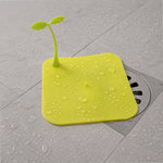 A bright yellow square silicone drain cover with a green sprout-like handle, covered in water droplets on a wet floor.