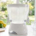 An empty, clear plastic dual-compartment beverage dispenser with a white base and two taps sits on a marble countertop with