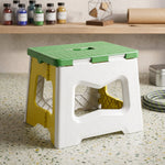 A green, white, and yellow plastic folding stool sits on a speckled counter with various craft supplies in the background.