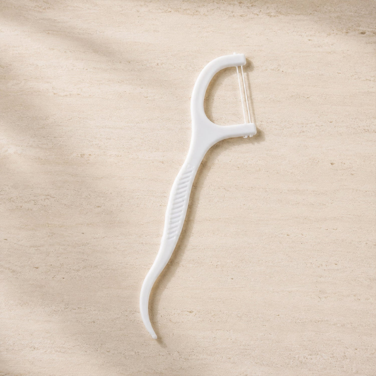 A single white dental floss pick with a textured handle lies on a light brown, textured surface.