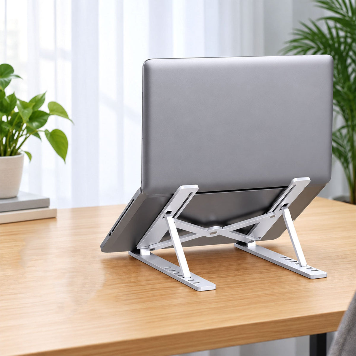 Silver laptop on a white adjustable stand, placed on a light wooden desk with a green plant nearby.