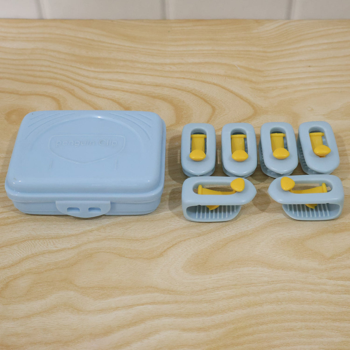 A light blue plastic storage case and six blue and yellow plastic clips are arranged on a light wooden surface.