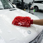 A hand wearing a red microfiber chenille wash glove cleans the sudsy white hood of a car in a garage setting.