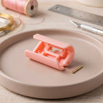 An open pink plastic bed sheet clip with a small gold pin rests on a pink plate, with craft supplies in the background.