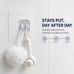 A white bath pouf and a white facial brush hang from a clear adhesive hook on a white tiled bathroom wall.