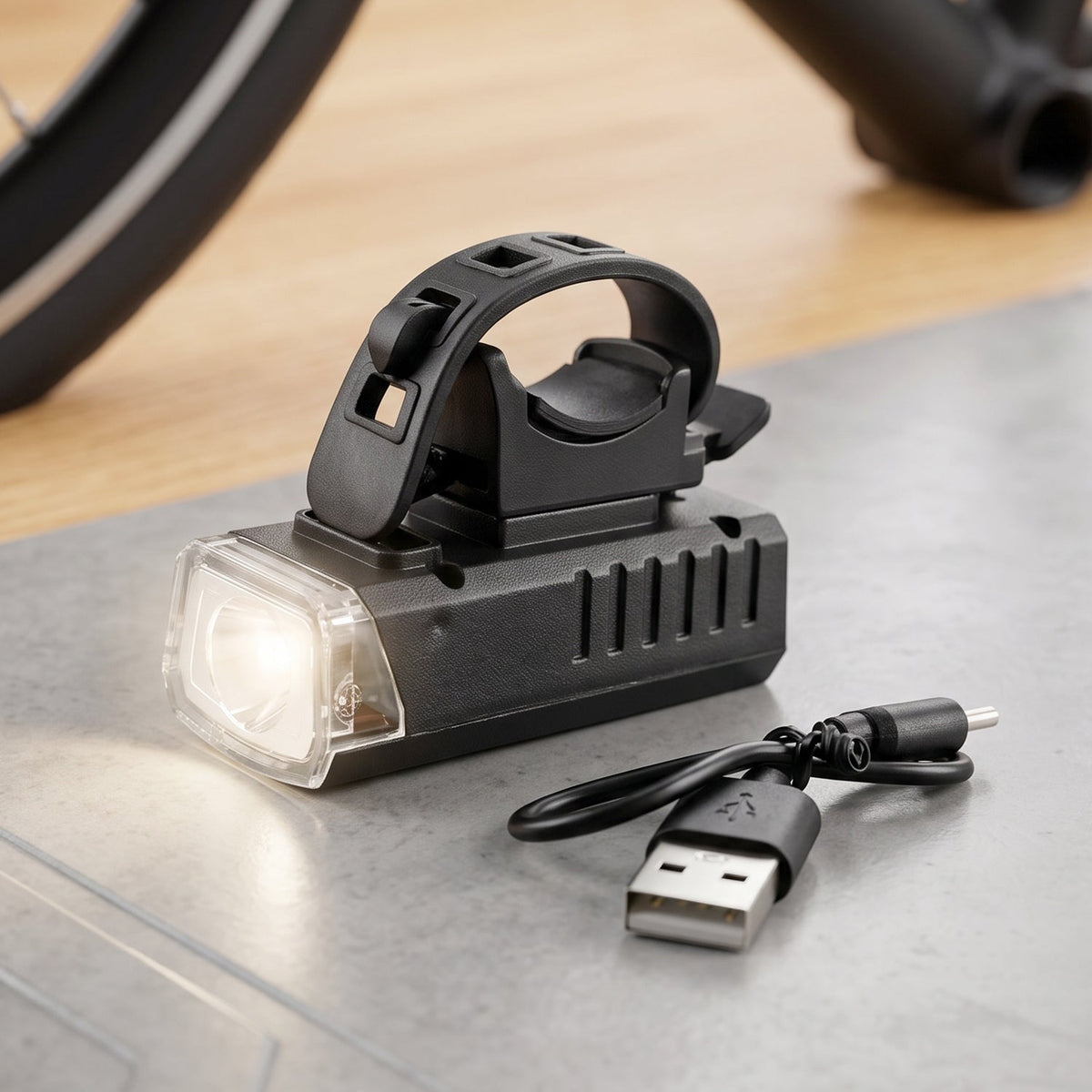 Black bicycle headlight with a clear lens and a USB charging cable on a gray surface, with a bicycle wheel in the background.