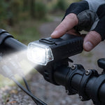 A gloved hand presses the button on a black bicycle headlight mounted on handlebars, emitting a bright beam.