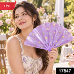 A woman smiles while holding a purple fan with cat and bow designs in a sunny outdoor garden.