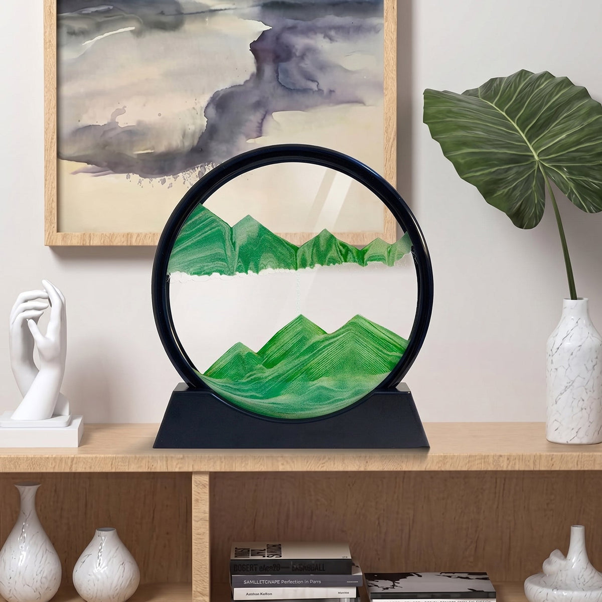 A black-framed circular sand art display with green sand forming mountains, on a light wooden shelf with decor.