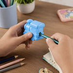 Hands sharpening a green pencil with a light blue train-shaped sharpener on a wooden desk with colored pencils.