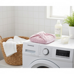 A pink microfiber hair towel wrap, folded into a square, sits on top of a washing machine next to a laundry basket and