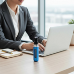 A blue refillable perfume bottle sits on a light wooden desk next to a laptop and notebook, with a person working in the