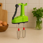 A green manual hand blender with black handle and red-tipped whisks sits on a kitchen counter with eggs and parsley in the