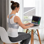 Woman wearing a grey posture corrector, typing on a laptop at a white table, with a window in the background.