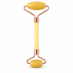 A yellow jade facial roller with rose gold colored metal accents is centered on a plain white background.