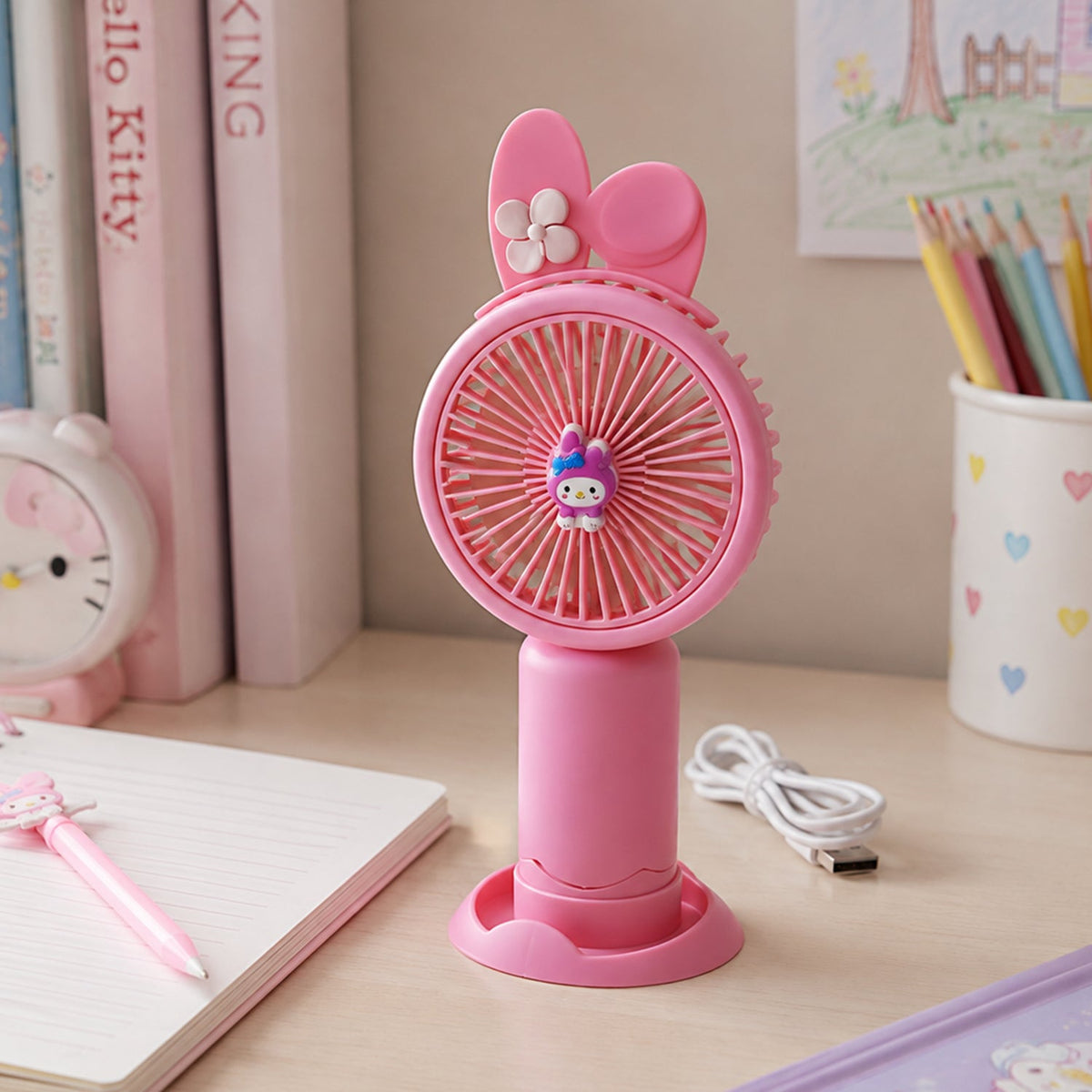A pink bunny ear mini handheld fan with a small character on the grill stands upright, with its USB cable on a white