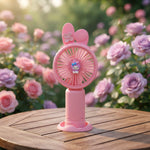 A young girl with flowing hair smiles as she holds a pink bunny ear mini handheld fan to her face, feeling the breeze.