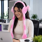 A young woman smiling and working on a laptop while wearing pink and white plush bunny earmuffs with long tails in an office