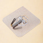 A close-up, angled view of a transparent self-adhesive hook with a silver metal hook on a light brown surface.