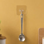 A silver spoon hanging from a transparent self-adhesive hook on a yellow wall, with a plant and wooden board visible.