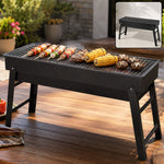 Black portable charcoal grill with food cooking on the grate, set outdoors on a wooden table.