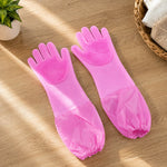 A pair of pink silicone scrubbing gloves with long cuffs on a wooden table next to a plant and a basket.