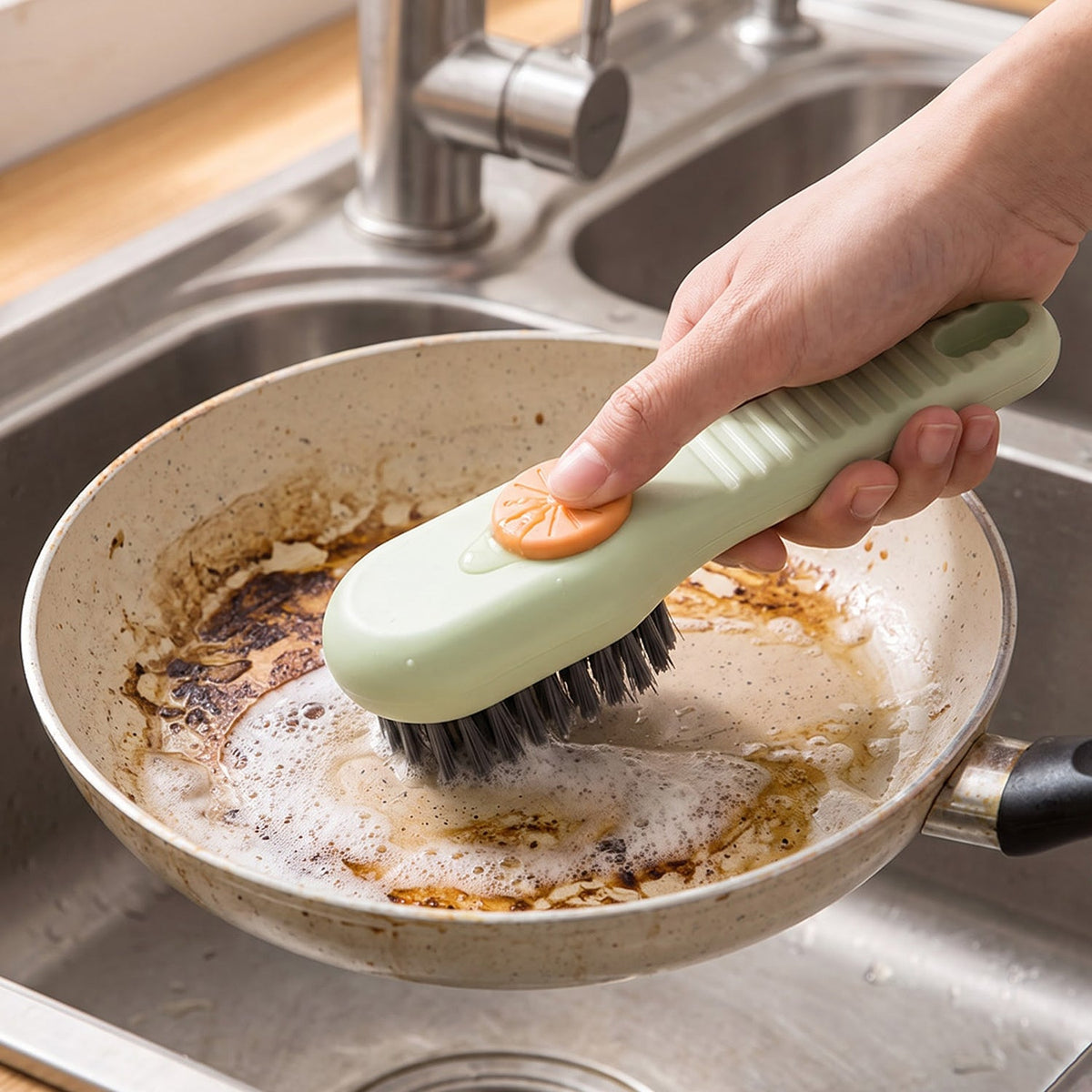 A hand uses a green and orange liquid-dispensing brush to clean a dirty frying pan in a stainless steel sink.