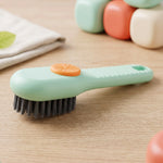 A green and orange liquid-dispensing brush rests on a wooden surface, with blurred colorful containers in the background.