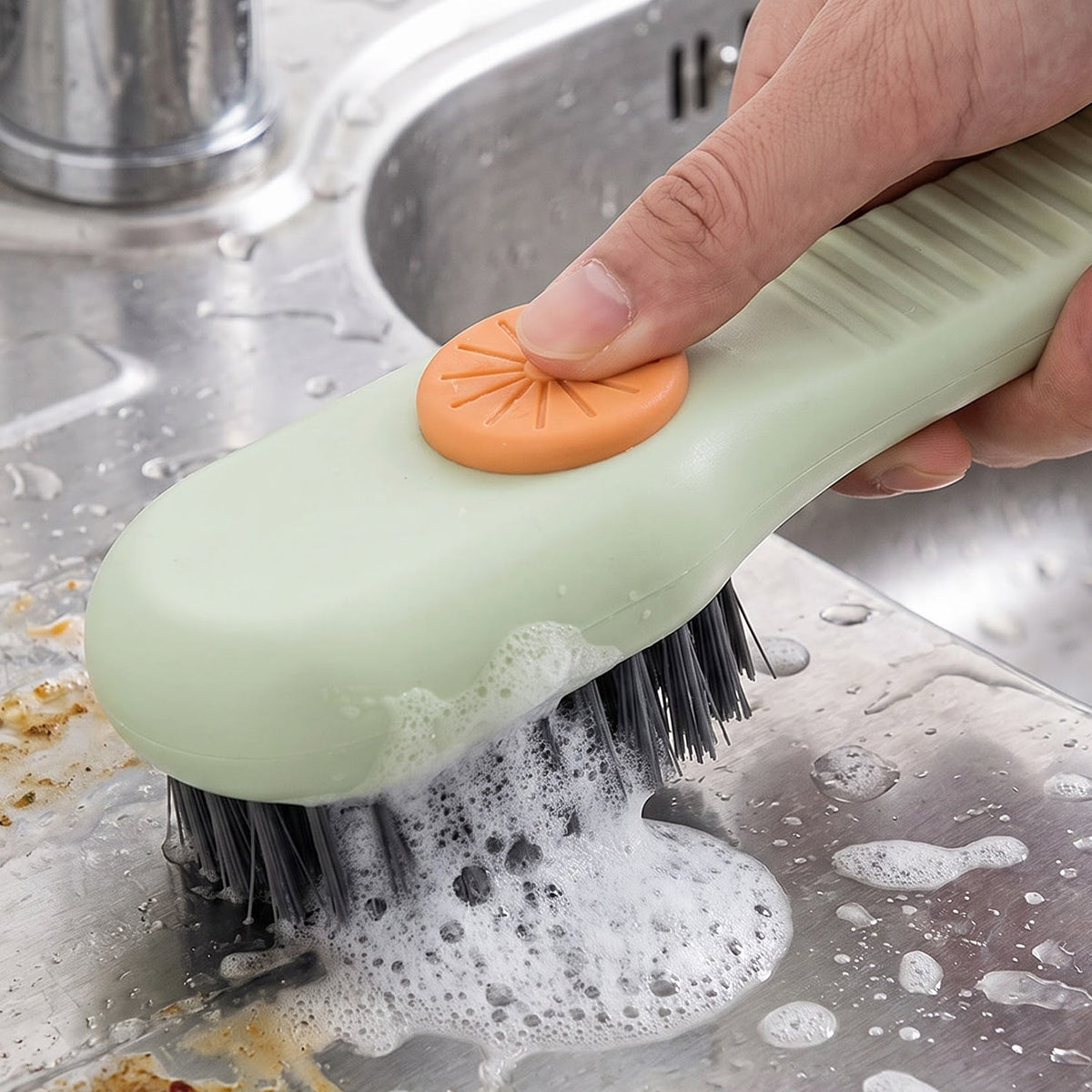 A hand uses a green and orange liquid-dispensing brush to scrub a stainless steel sink, creating suds.