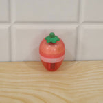 A pink and red strawberry-shaped sharpener with a green stem rests on a light wooden surface against white tiles.