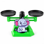 A green plastic elephant-themed math balance scale with two black weighing pans on a white background.