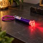A purple laser pointer keychain with a clear flower cap illuminates a red light pattern on a dark surface.
