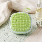 A light green square silicone scalp massage brush with numerous soft bristles lies on a speckled green surface next to a