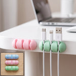 A pink and a green silicone cable organizer are attached to a white desk, holding three charging cables.
