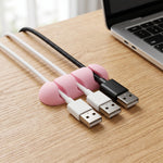 A pink silicone cable organizer is attached to a wooden desk, holding three charging cables next to a laptop.