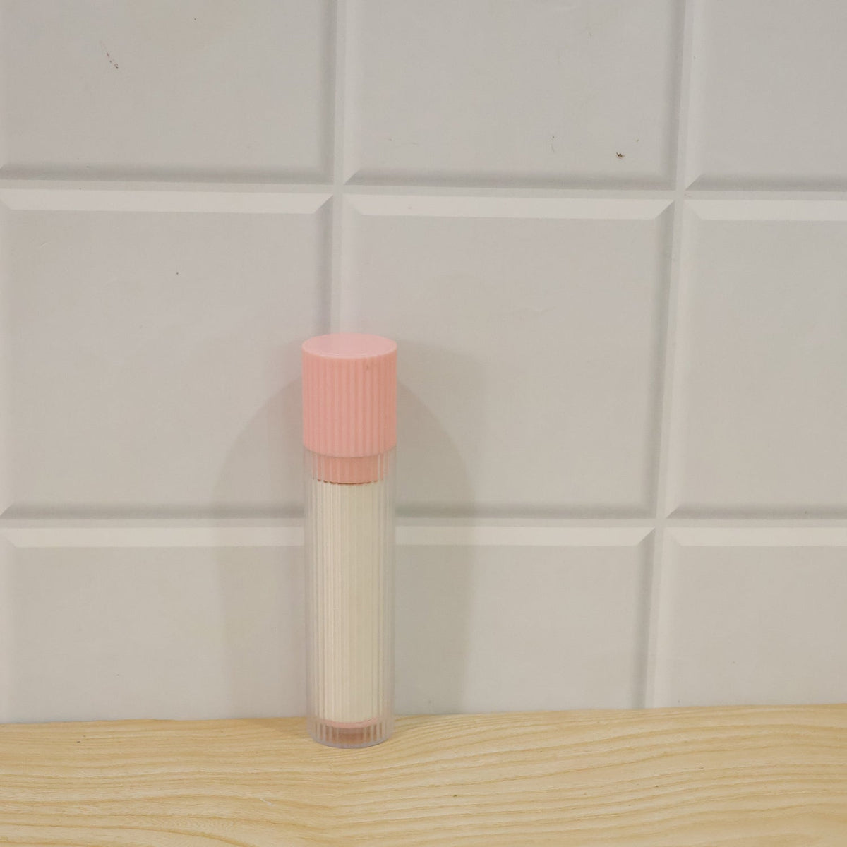 A pink and white lint roller with its clear ribbed protective case stands upright on a wooden surface against a white tiled