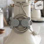 A gray silicone cord organizer with two smiling faces, holding a coiled light gray appliance cord on a white stand – view 3