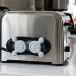 A gray silicone cord organizer with two smiling faces, holding a coiled black appliance cord on a stainless steel toaster.